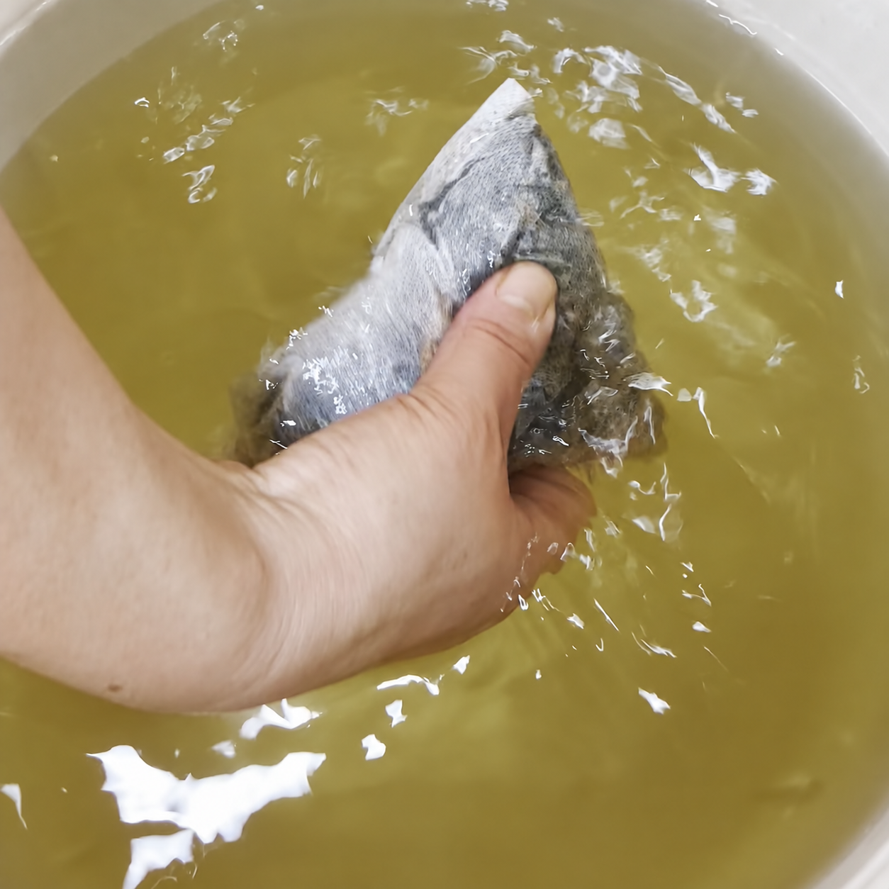AI-edited prototype illustration based on a video frame: a hand holding a filled cloth sachet in botanical bathwater; basin color adjusted.